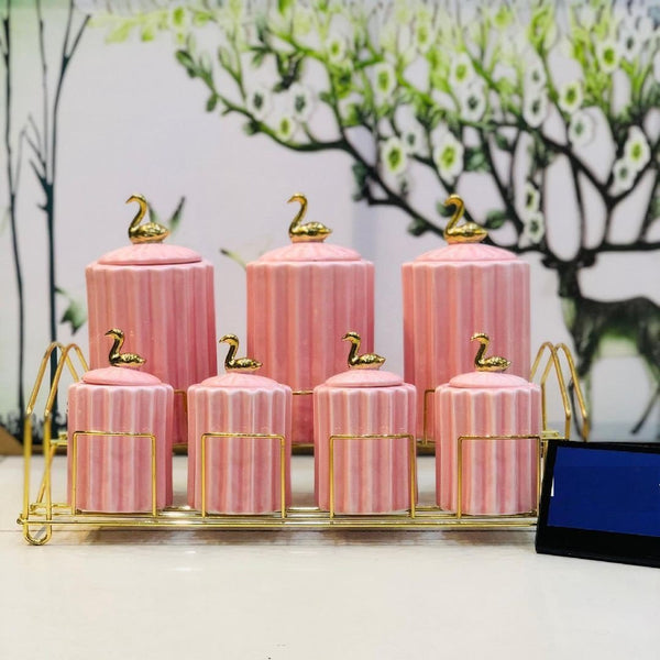 7-piece pink ceramic spice jar set with golden stand on a table top, with lids featuring a small gold swan design.