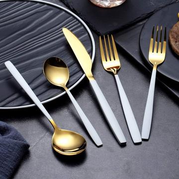 A complete cutlery set featuring gold and white-handled spoons, forks, knives, and teaspoons elegantly placed on black plates, set against a sleek black tablecloth. The clean, sophisticated setup showcases each piece of the set for an upscale dining experience.