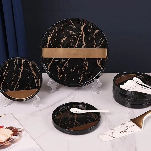 Black and gold ceramic dessert set displayed on a tabletop, featuring one large plate with six smaller plates stacked, a spatula and spoons.