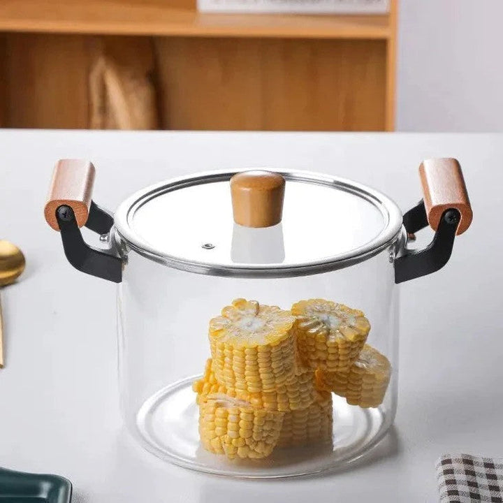 Glass cooking pot on a tabletop filled with corn, highlighting the wooden handle and clear glass lid.