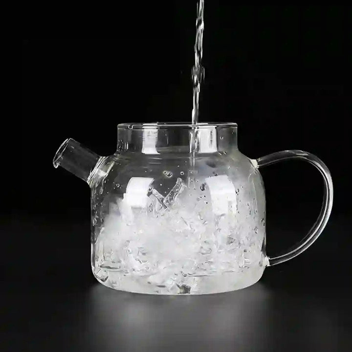 Glass kettle filled with ice, as water is poured in, emphasizing its versatility for serving cold beverages in addition to tea.