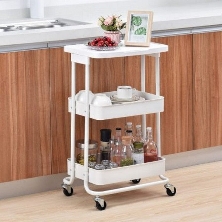 White 3-tier metal storage trolley for kitchen organization, with decorative items on top shelf, mugs and cups on middle shelf, and spice jars on bottom shelf.