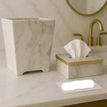 Marble-patterned tissue box and trash can on a bathroom counter with a mirror and faucet in the background.