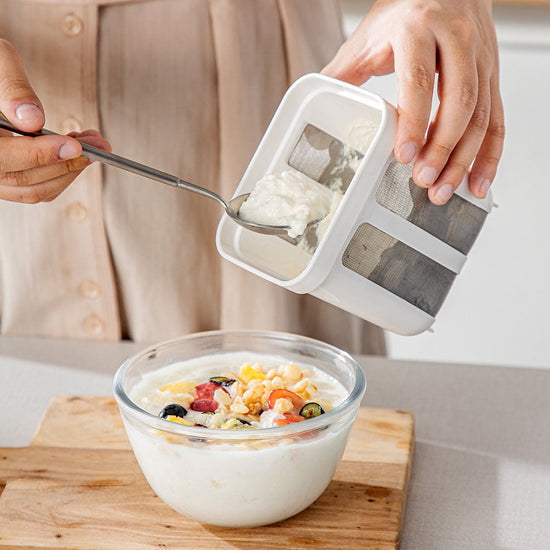 Person using a yogurt maker with a bowl of yogurt and fruit on a wooden cutting board.