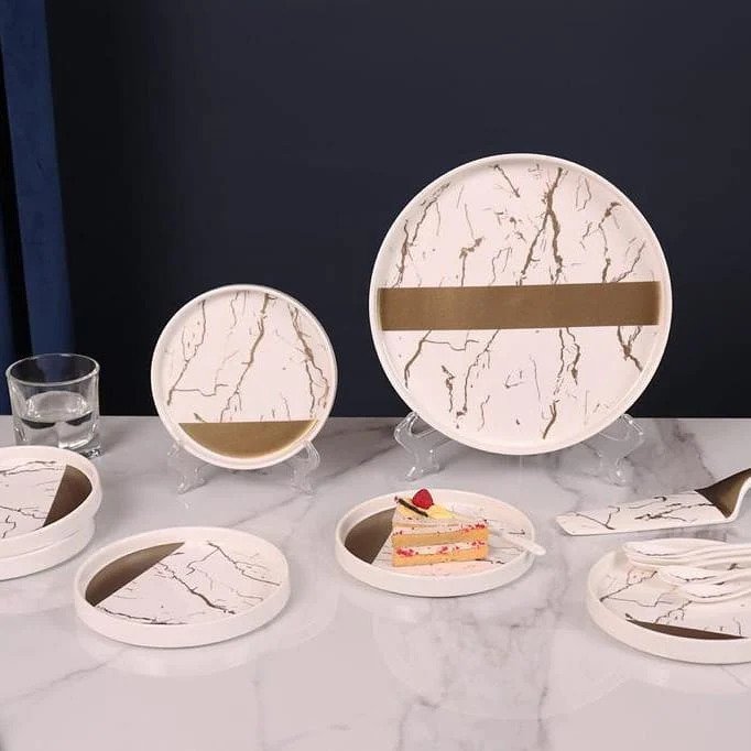 white and gold ceramic dessert set displayed on a tabletop, featuring one large plate with six smaller plates stacked, a spatula and spoons.